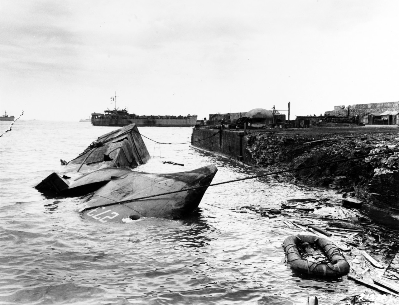Anzio Campaign, 1944. HMS LCI-273 sunk at Anzio, March 28, 1944, soon after she was hit by a German air attack. Note ship's hull broken in half, damage to pier alongside, life raft floating amid debris and pillbox at the end of the pier. Photographed by Leibowitz